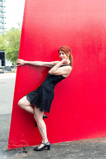 Stylish mature woman in a black dress and heels poses playfully against a vibrant red wall, creating a striking contrast in an urban environment