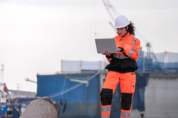 istock Two port engineers in high-visibility orange uniforms and hard hats communicate while inspecting dockside logistics using a laptop and walkie-talkie, symbolizing teamwork and safety. 2223286732