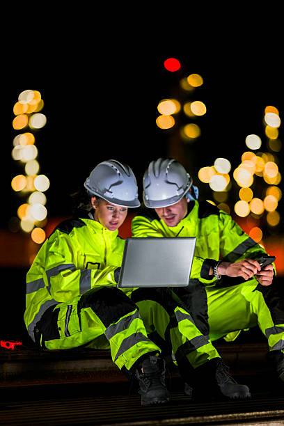 istock Two industrial workers in high-visibility gear and helmets engage in a serious discussion while using a laptop and smartphone under night lighting at a worksite. 2223286819