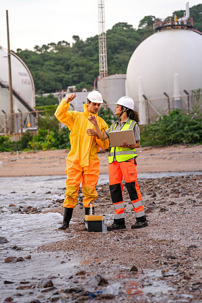 istock Two engineers in safety gear collect water samples and input data on a laptop near coastal chemical storage tanks. Field research and pollution monitoring in action. 2223288020