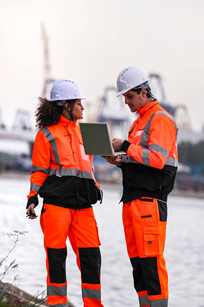 istock Two port engineers in high-visibility orange uniforms and hard hats communicate while inspecting dockside logistics using a laptop and walkie-talkie, symbolizing teamwork and safety. 2223288835