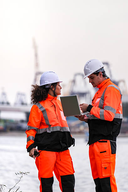 istock Two port engineers in high-visibility orange uniforms and hard hats communicate while inspecting dockside logistics using a laptop and walkie-talkie, symbolizing teamwork and safety. 2223288964