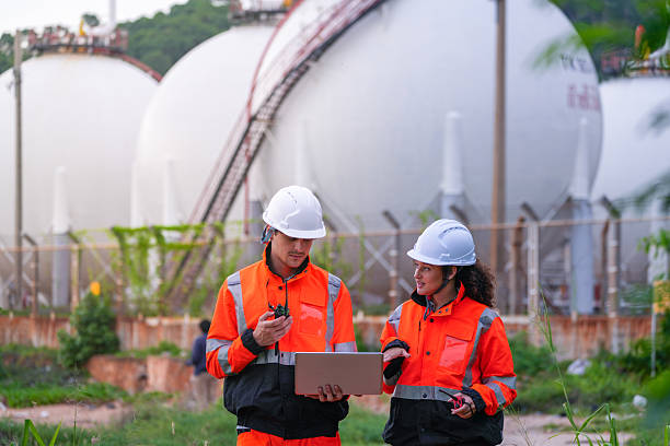 istock Two engineers in high-visibility safety gear and hard hats discuss project plans on a laptop in front of large spherical gas storage tanks at an industrial coastal facility. 2223289026