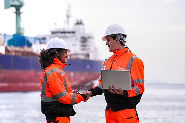 istock Two port engineers in high-visibility orange uniforms and hard hats communicate while inspecting dockside logistics using a laptop and walkie-talkie, symbolizing teamwork and safety. 2223289072
