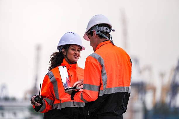 istock Two port engineers in high-visibility orange uniforms and hard hats communicate while inspecting dockside logistics using a laptop and walkie-talkie, symbolizing teamwork and safety. 2223289117