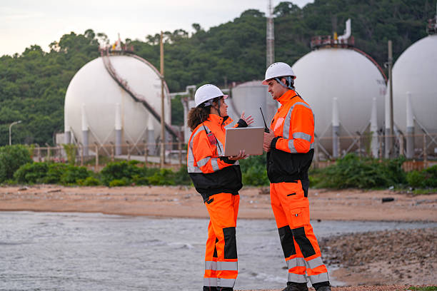 istock Two engineers in high-visibility safety gear and hard hats discuss project plans on a laptop in front of large spherical gas storage tanks at an industrial coastal facility. 2223289239