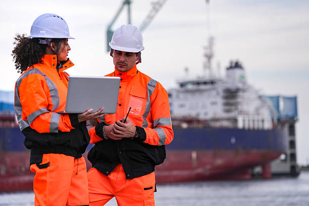 istock Two port engineers in high-visibility orange uniforms and hard hats communicate while inspecting dockside logistics using a laptop and walkie-talkie, symbolizing teamwork and safety. 2223289363