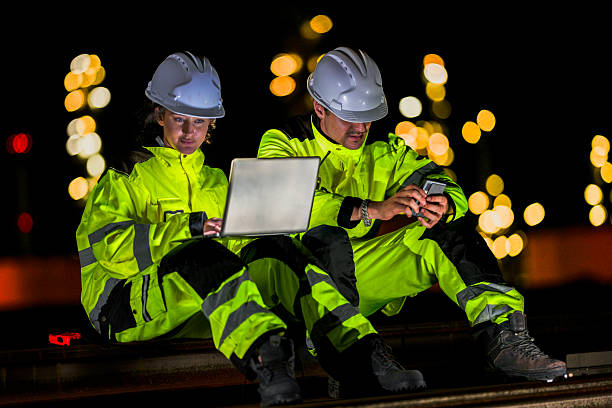 istock Two industrial workers in high-visibility gear and helmets engage in a serious discussion while using a laptop and smartphone under night lighting at a worksite. 2223290058