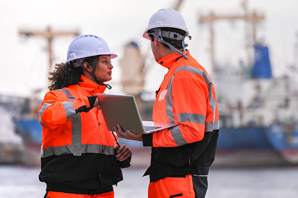istock Two port engineers in high-visibility orange uniforms and hard hats communicate while inspecting dockside logistics using a laptop and walkie-talkie, symbolizing teamwork and safety. 2223290093