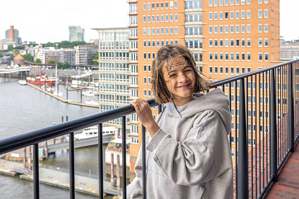 A smiling girl in a grey hoodie stands on a balcony with city buildings and a river marina behind her. A joyful moment in an urban setting.