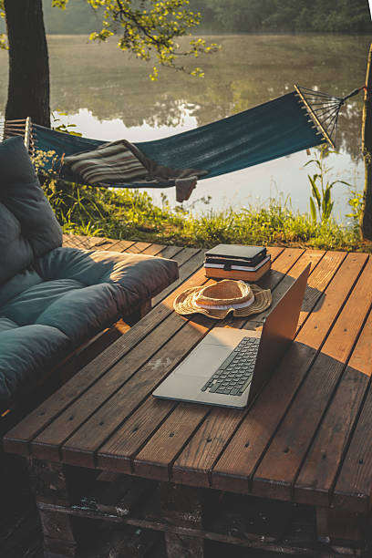 Laptop placed on a wooden table in a tranquil forest setting with a hammock and soft morning light, ideal for remote work or digital nomad lifestyle.