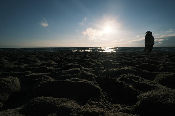 Dramatic lighting as a lone figure stands by the sea, captured from the beach’s sand. The low sun creates striking shadows.