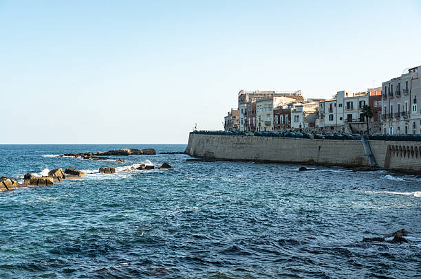 Syracuse, Italy, November 1, 2024: Stone coastal wall and buildings along Ortigia waterfront in Syracuse. Urban shoreline with masonry structures and waves breaking on rocks, editorial image