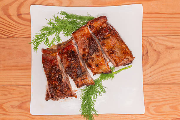 Serving of the sliced pork ribs, baked in special sauce, decorated with fresh dill twigs on a white square plate on a wooden rustic table, top view
