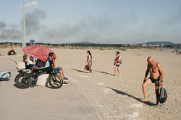 July 2025, Italy – People enjoy a summer day on a public beach. A man sits under an umbrella next to an e-bike, while others walk on the sand or prepare to enter the water. In the background, a column of smoke can be seen, probably caused by a forest fire.