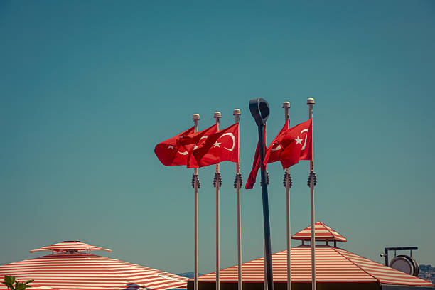 Multiple Turkish flags waving in the wind against a vivid blue sky in Istanbul, Türkiye. The red flags with the white star and crescent symbol represent national pride and identity. In the foreground, striped parasols enhance the vibrant summer mood. This image evokes a sense of patriotism, Mediterranean lifestyle, and travel atmosphere, ideal for tourism, culture, or seasonal editorial content.