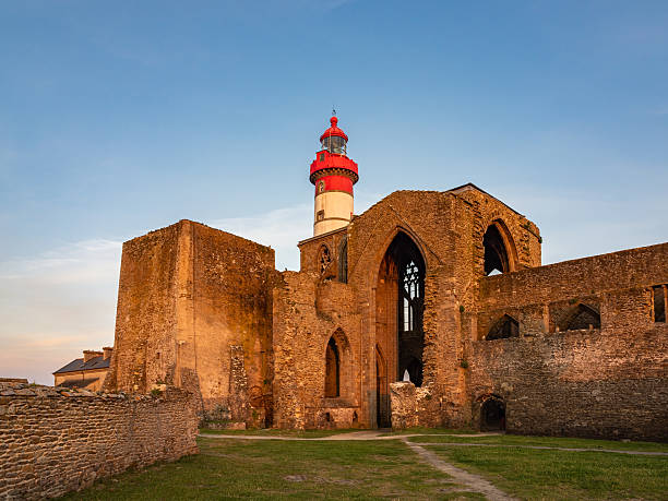 A dramatic blue-hour scene of the Kermorvan Lighthouse standing on a rocky headland in Le Conquet, Brittany, France. The lighthouse rises above turbulent Atlantic waters, connected by a historic stone path that curves over dark, jagged rocks. Heavy clouds sweep across the sky, enhancing the moody coastal atmosphere. The rugged textures, shifting tides, and remote maritime setting emphasize the lighthouse’s enduring role as a beacon of navigation and safety on one of France’s wildest shorelines