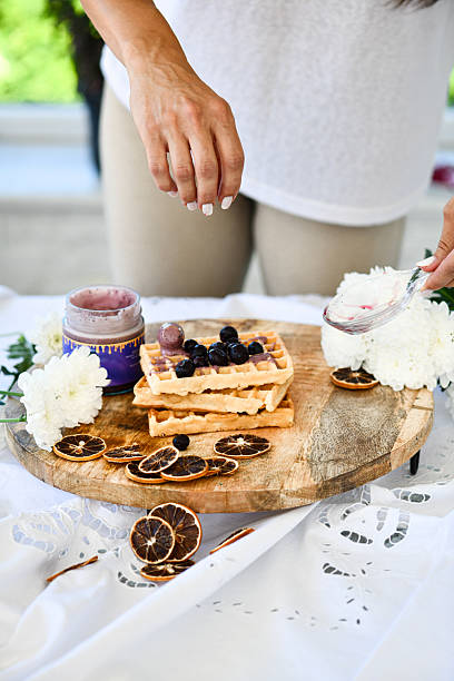 Female Gently Adding Fresh Blueberries On Top Of Jam-Covered Waffles Placed On Wooden Board