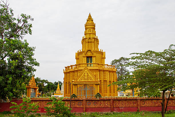 A radiant golden pagoda, possibly in Cambodia, rises majestically among lush green trees beneath a bright sky. The shimmering structure contrasts beautifully with its natural surroundings, evoking a sense of serenity and spiritual splendor.