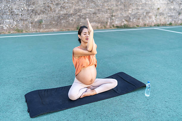 Pregnant woman in her 30s practicing yoga on a mat outdoors, seated in a meditative pose, focusing on mindfulness and prenatal well-being in a serene setting