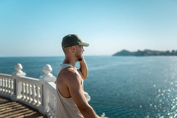 Young man in casual attire stands on a balcony overlooking a vast ocean, enjoying the tranquil scenery under a clear blue sky. The setting exudes relaxation and freedom