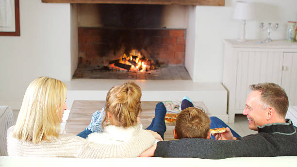 istock Family Sitting On Sofa In Lounge Next To Open Fire Eating Pizza 2223400726