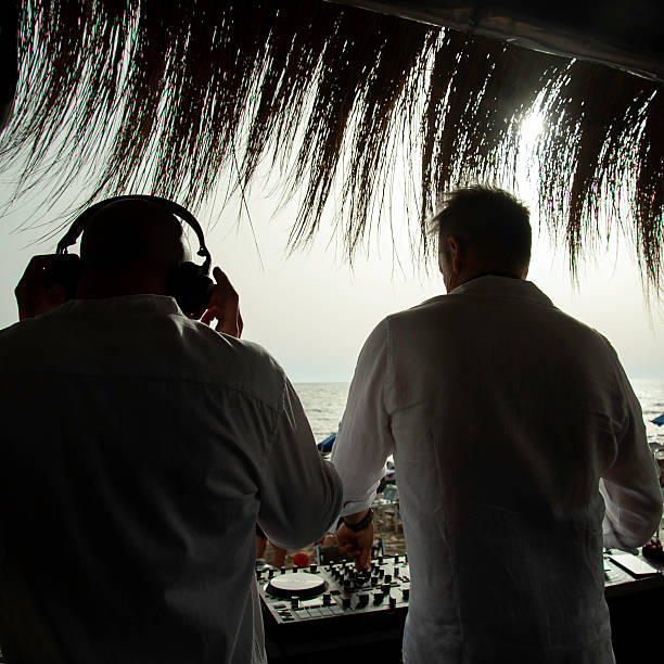 Two DJs performing at a beachside bar under a thatched roof with ocean view and sunset light. Backlit scene with music, summer vibe, and party atmosphere. Entertainment and lifestyle concept.