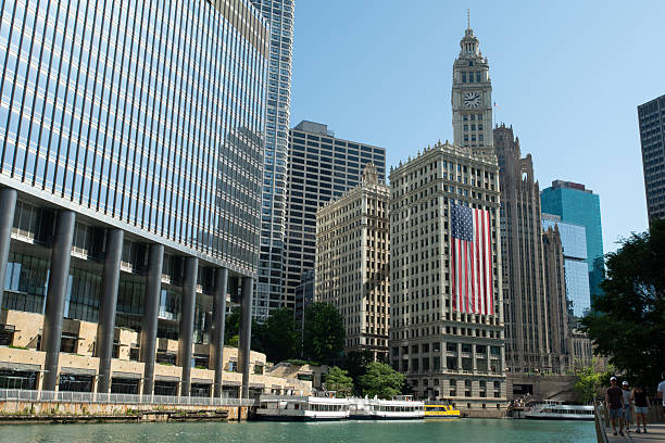 Tourists (right) walk along the Chicago Riverwalk and take in a Wrigley Building and Trump Tower view (left). Also visible are numerous Chicago water taxis (bottom)
