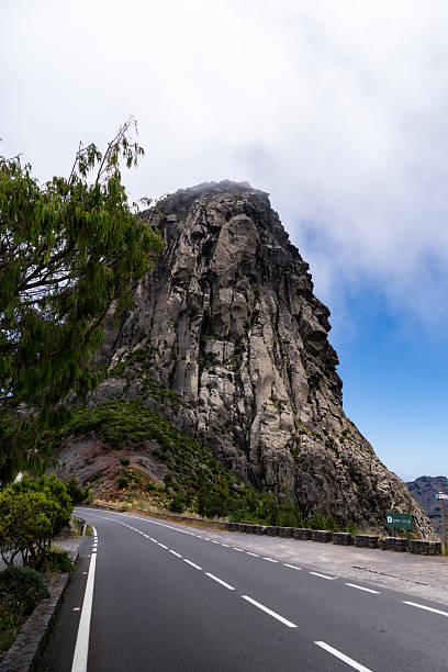 Towering mountain cliff rises beside an empty road under a partially cloudy sky, creating a dramatic natural landscape
