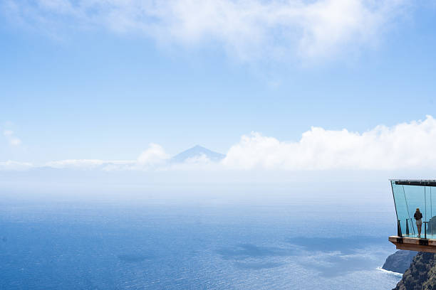 Stunning glass balcony juts out over a cliff, offering breathtaking views of the ocean and distant mountains under a clear blue sky