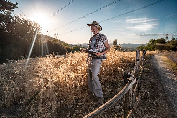 istock Farmer Examining Crops in Rural Field During Sunset with a Notebook and Hat 2223429387