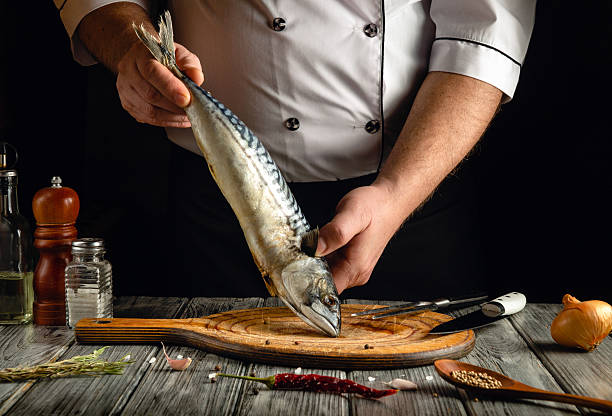 istock A chef holds a fresh mackerel above a wooden cutting board in a cozy kitchen. Various spices, herbs, and ingredients surround the preparation area, enhancing the culinary atmosphere 2223441784