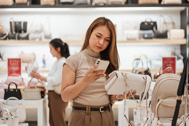 Asian woman comparing prices or reading online reviews while shopping for a handbag. Using smartphone in-store. Concept of smart consumer lifestyle.