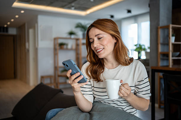 istock Smiling woman using smartphone and drinking coffee on sofa at home 2223451877