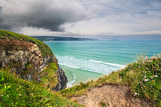 From a rugged cliff edge, the viewer takes in the dramatic coastal scene: vibrant turquoise waves crash below, contrasting with the moody, cloudy sky overhead. In the distance, a coastal town stretches along the shore, adding a touch of human presence to the wild landscape. This image captures the raw beauty and changing atmosphere of the coast.