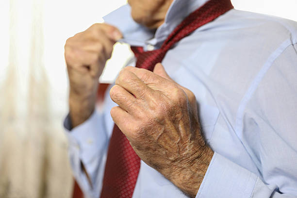 Detailed close-up of elderly male hands adjusting a red polka-dot necktie while wearing a light blue dress shirt. The image focuses on texture, age, and personal grooming, suggesting themes of dignity, routine, and daily preparation.