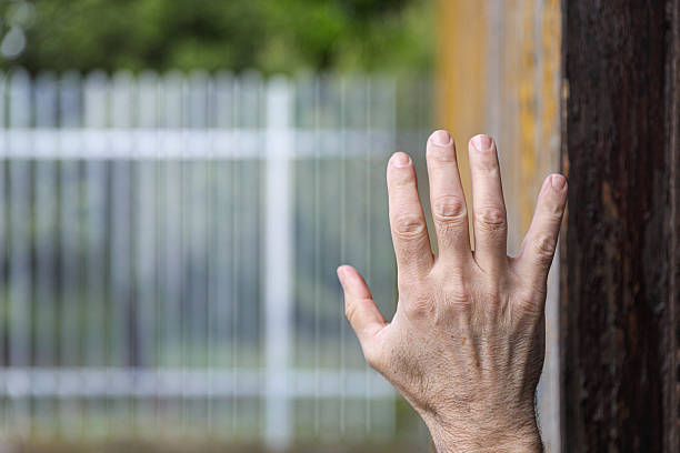Close-up of an open hand reaching toward a blurred metal fence, symbolizing longing, freedom, or the emotional boundary between confinement and escape. The contrast between the soft background and focused hand conveys a message of hope, waiting, or the desire to overcome barriers — physical or emotional.
