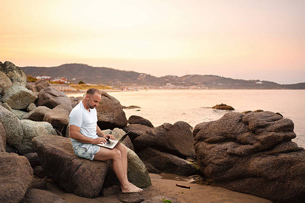 istock Man working on laptop sitting on rock at beach during sunset 2223461370