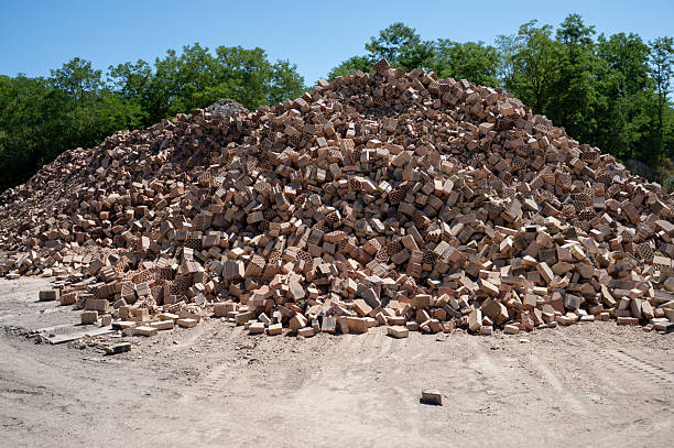 A towering pile of mixed clay bricks is shown under bright sunlight in an outdoor industrial setting, with green trees in the background. The bricks vary in shape and structure, contributing to a rugged and textured visual. The image reflects mass production, material accumulation, and construction industry scale.