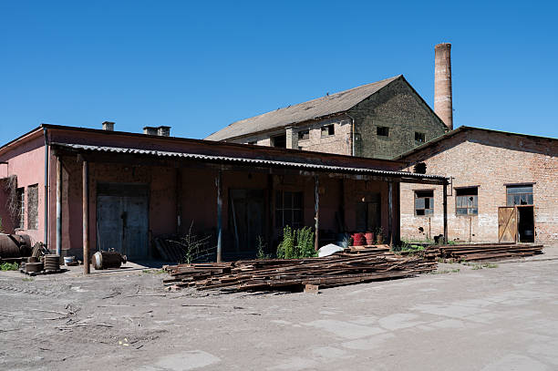 An abandoned industrial brick factory complex under a clear blue sky, featuring weathered red brick walls, a rusted metal roof, and a tall chimney stack. Metal pipes and construction debris are scattered in the courtyard, adding to the sense of decay and industrial history. The scene evokes a mood of abandonment and past productivity.