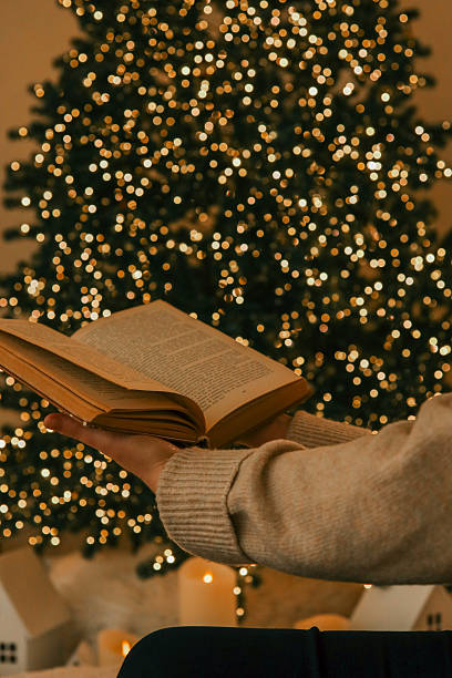 istock Person holding an open book in front of a decorated Christmas tree with glowing lights. A cozy setting featuring warm tones, miniature houses, and candles, ideal for holiday reading moments 2223480077