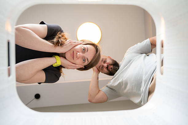 A couple observes an empty laundry hamper from an unusual perspective, expressing curiosity and surprise. The image embodies a unique angle, modern domesticity, and emotions of intrigue and puzzlement.