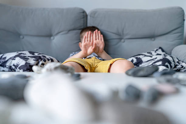 A young child sits on a comfortable sofa, playfully covering their face with hands, expressing emotions or shyness. The blurred rocks collection on the table on foreground adds an artistic touch, highlighting the child and the cozy setting.