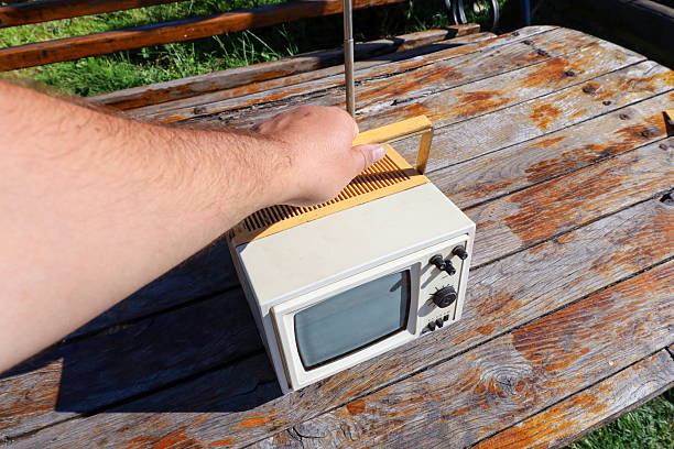 A portable retro television with a retractable antenna stands on a wooden table outdoors. A hand holds it by the handle, emphasizing the device's mobility. This nostalgic image evokes memories of the past.