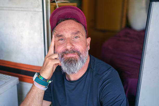 Close-up of a mature man with a beard and a cap, sitting on the doorstep of his camper van, looking at the camera