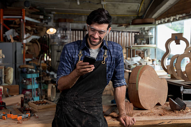 Happy carpenter taking a break in woodworking shop while checking smartphone. Wearing safety goggles and apron, standing near wooden instrument. Concept of craft, tech use, and modern labor.