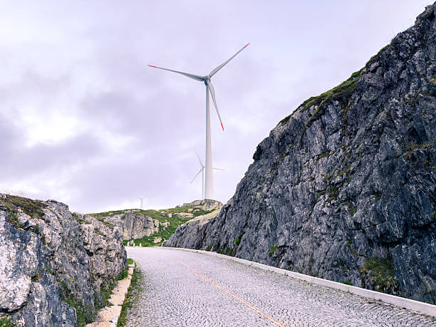 Tall wind turbines stand on green hills beside a winding cobblestone road through rocky terrain. Renewable energy structures in a natural mountainous landscape. Sustainable power and nature.