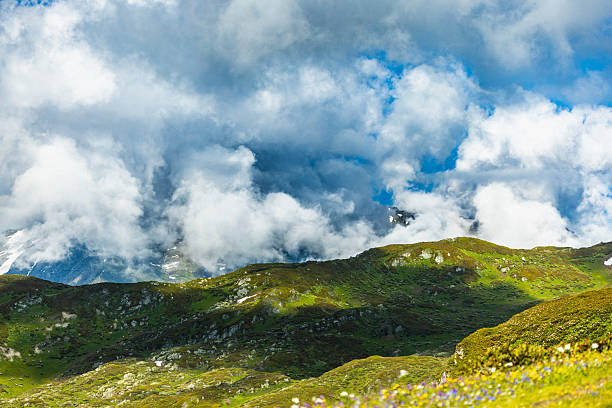 Beautiful mountain scenery featuring snow-capped peaks, vibrant green foothills, and dynamic clouds under a blue sky, evoking tranquility and nature’s grandeur.