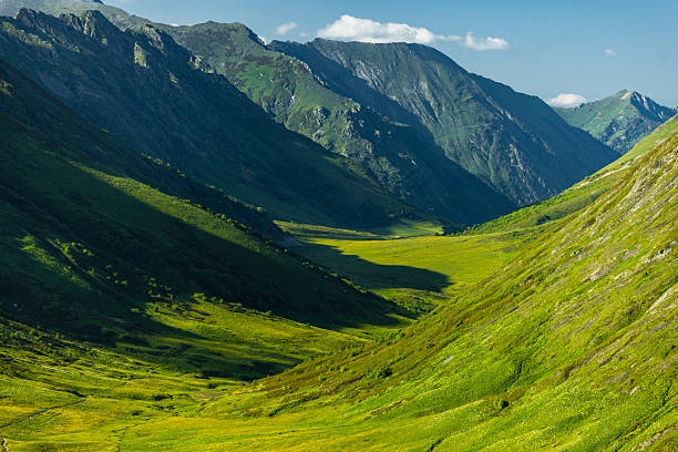 A vibrant image showcasing a valley drenched in sunlight, with tents scattered on the green terrain, surrounded by towering mountains casting shadows.