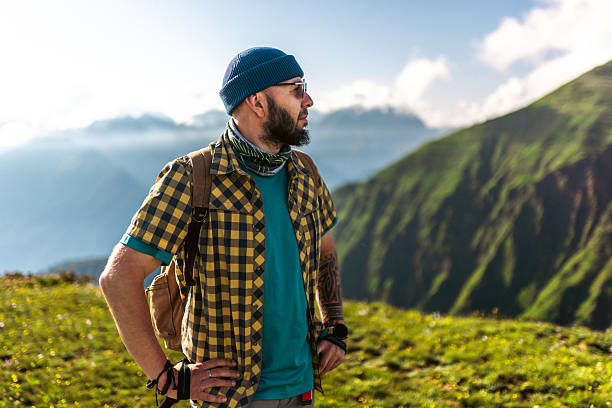 A bearded man in casual hiking attire uses trekking poles while enjoying a sunny mountain trek. The scene highlights the beauty of outdoor activities, surrounded by green hills and an inspiring open sky.